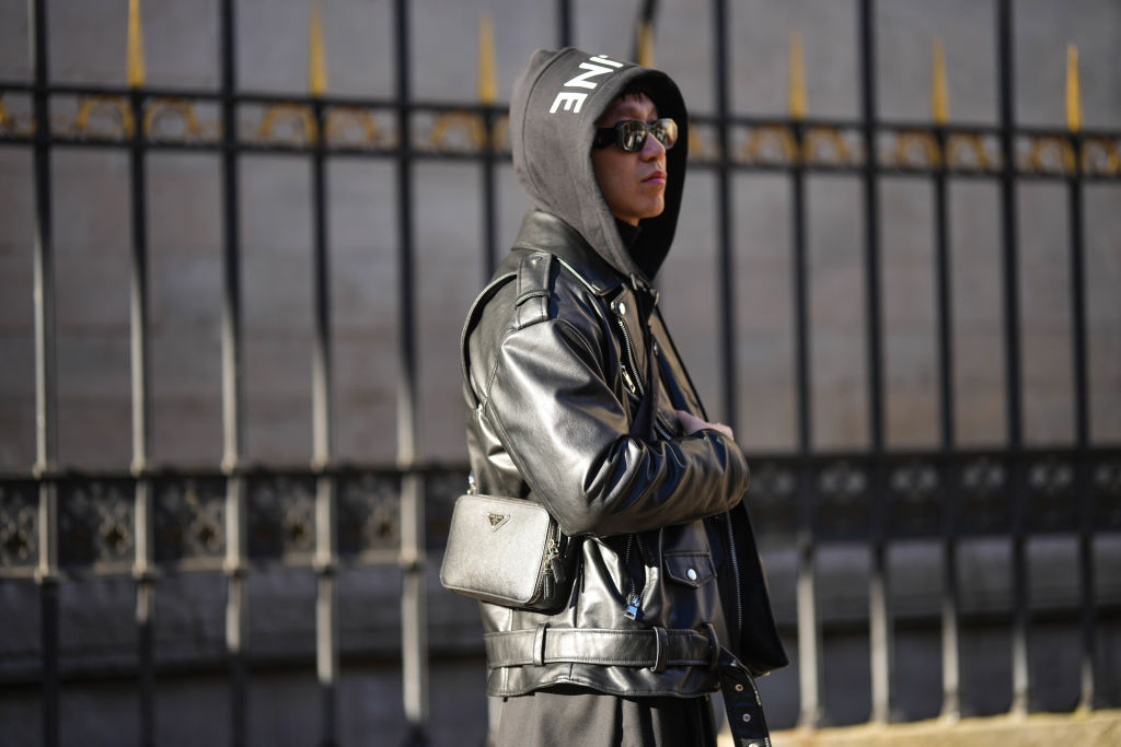 A guest wears black sunglasses, a black hoodie sweater with white logo print pattern from Celine, a black shiny leather zipper biker jacket, a black long coat, a black shiny grained leather crossbody bag from Prada, outside BlueMarble, during Paris Fashion Week