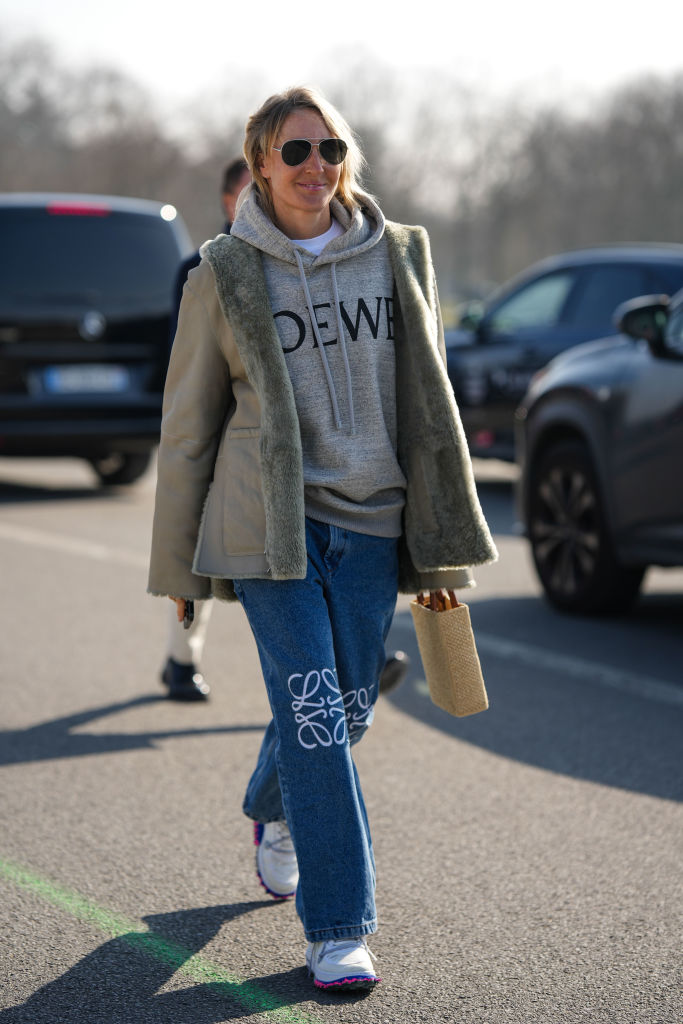 A guest wears black aviator sunglasses, a white t-shirt, a pale gray with embroidered black logo pattern hoodie sweater from Loewe, a khaki suede and fur interior jacket, blue denim with embroidered white knees pattern large denim pants, a beige braided wicker and black logo handbag from Loewe, white sneakers , outside Loewe, during Paris Fashion Week