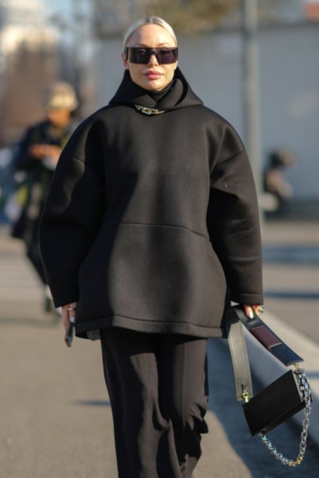 MILAN, ITALY - JANUARY 16: A guest wears black futurist sunglasses, a large silver chain necklace, a black hoodie oversized nylon sweater, black large flared pants, a black shiny leather handbag from Prada, silver rings, black shiny leather ankle boots, outside the Prada fashion show during the Milan Men's Fashion Week - Fall/Winter 2022/2023 on January 16, 2022 in Milan, Italy. (Photo by Edward Berthelot/Getty Images)