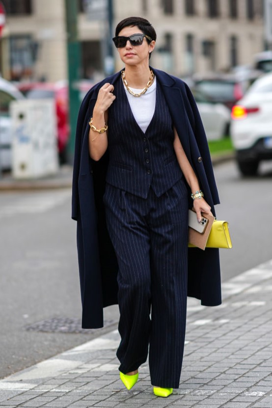 MILAN, ITALY - FEBRUARY 24: Chrisa Pappas wears black sunglasses, a gold large necklace, a matching gold large chain bracelet, a gold watch, a silver and blue pearls bracelet, a yellow shiny leather Kelly clutch from Hermes, a white t-shirt, a navy blue striped print pattern sleeveless gilet, matching navy blue striped print pattern large suit pants, a navy blue long coat, neon yellow shiny leather block heels pointed shoes, outside the Max Mara fashion show, during the Milan Fashion Week Fall/Winter 2022/2023 on February 24, 2022 in Milan, Italy. (Photo by Edward Berthelot/Getty Images)