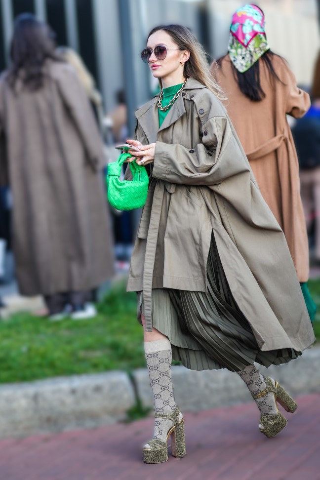 MILAN, ITALY - FEBRUARY 25: A guest wears brown sunglasses, gold earrings, a gold large chain necklace, a dark green high neck pullover, a brown oversized belted cloak coat, a khaki pleated / accordion midi skirt, beige with brown GG monogram print pattern socks, a green braided shiny leather Jodie handbag from Bottega Veneta, gold rings, a gold bracelet, khaki glitter / sequins platform high heels sandals , outside the Gucci fashion show, during the Milan Fashion Week Fall/Winter 2022/2023 on February 25, 2022 in Milan, Italy. (Photo by Edward Berthelot/Getty Images)