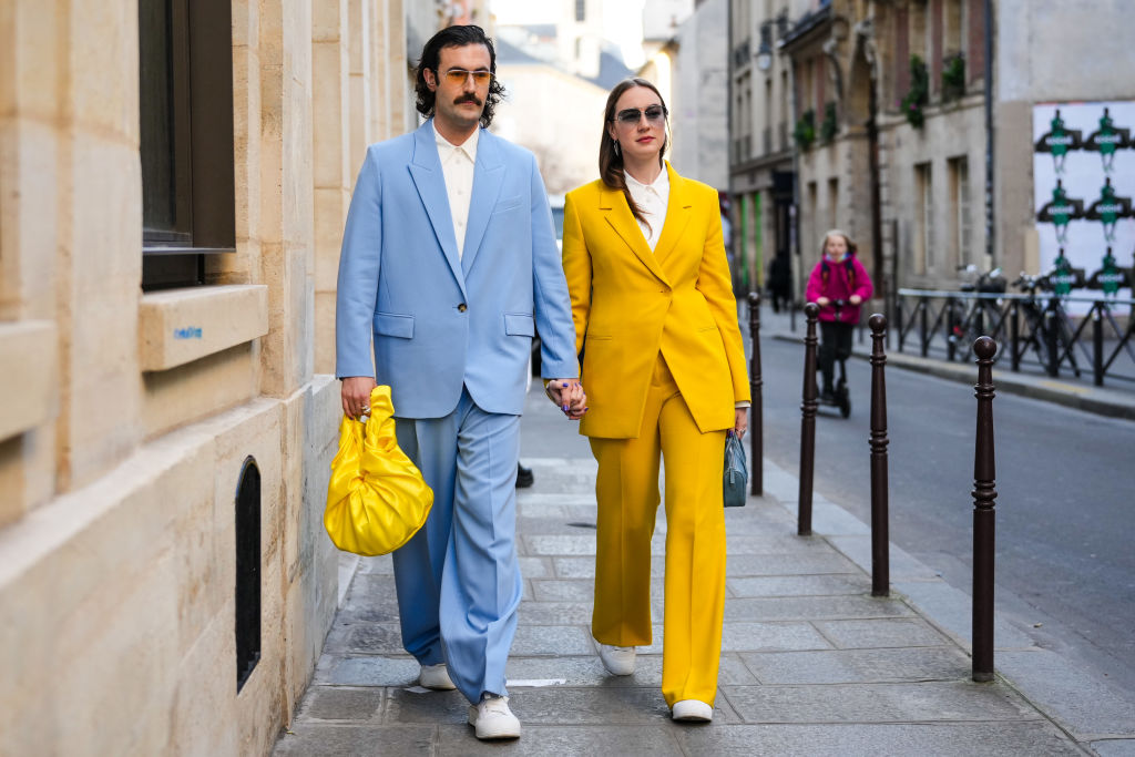 PARIS, FRANCE - FEBRUARY 28: A guest (L) wears sunglasses, a white shirt, a pale blue blazer jacket, matching pale blue suit pants, a yellow shiny satin ruffled handbag, white sneakers, a guest (R) wears sunglasses, a white shirt, a yellow blazer jacket, matching yellow suit pants, white sneakers, a gray shiny leather handbag, silver rings , outside Vaquera, during Paris Fashion Week - Womenswear F/W 2022-2023, on February 28, 2022 in Paris, France. (Photo by Edward Berthelot/Getty Images)