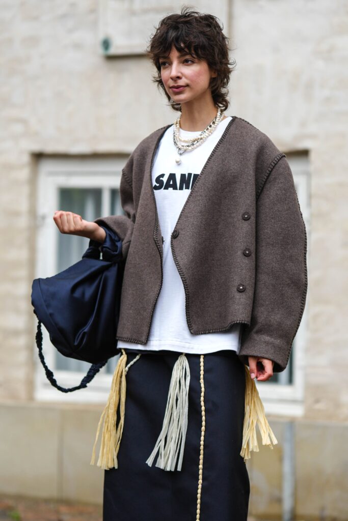 COPENHAGEN, DENMARK - AUGUST 09: A guest wears a silver large chain necklace, a white pearls necklace, a white with black logo print pattern t-shirt from Jil Sander, a dark gray oversized blazer jacket, a navy blue nylon large handbag, navy blue denim with embroidered yellow and pale gray fringed midi skirt, black shiny leather ballerinas , outside Marimekko, during the Copenhagen Fashion Week Spring/Summer 2024 on August 09, 2023 in Copenhagen, Denmark. (Photo by Edward Berthelot/Getty Images)