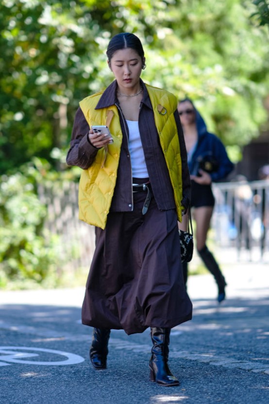 NEW YORK, NEW YORK - SEPTEMBER 08: A guest wears dark red brown jacket with yellow puffer jacket vest, white tank top, silver necklace, shiny black leather belt, dark red midi skirt, shiny black leather bag, shiny black heeled slouchy leather boots, outside Off-White, during the New York Fashion week Spring/Summer 2025 on September 08, 2024 in New York, New York.