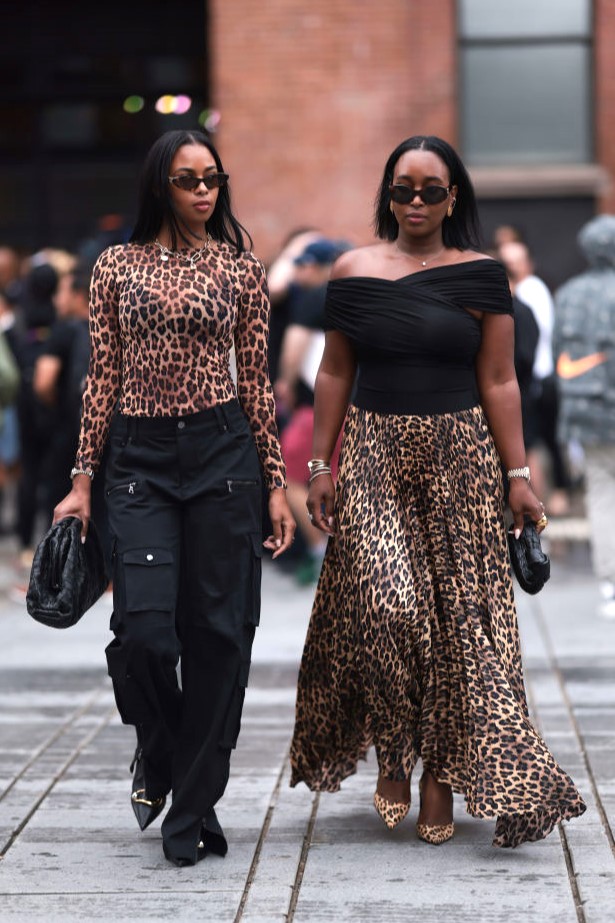 NEW YORK, NEW YORK - SEPTEMBER 07: A guest (L) wears Miu Miu brown sunglasses, gold pendant chain necklace, brown animal print / leopard print sheer long sleeve top, black cargo long pants with pockets, Bottega Veneta black woven leather pouch / clutch bag and black shiny leather pumps / heels; Next to her a guest (R) seen wearing Celine brown oval Triomphe sunglasses, Dior vintage gold earrings, diamond pendant necklace, black cotton off-shoulder top, brown animal print / leopard print flared pleated long skirt, Bottega Veneta black woven leather mini Jodie bag and animal print leather pumps / heels, outside the Alice + Olivia by Stacey Bendet Spring 2025 presentation, during the New York Fashion Week, on September 07, 2024 in New York City. (Photo by Jeremy Moeller/Getty Images)