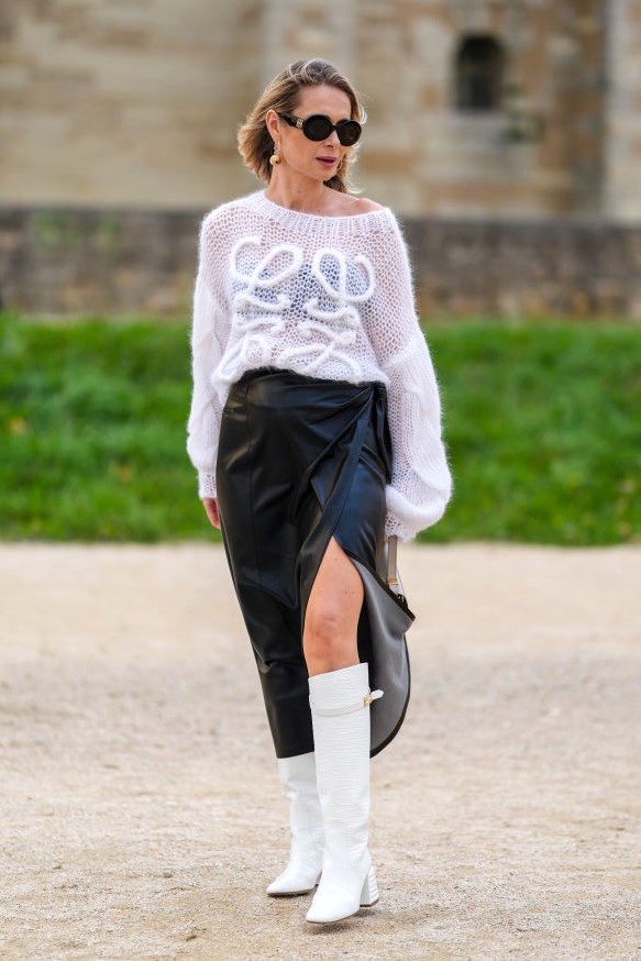 PARIS, FRANCE - SEPTEMBER 27: A guest wears black sunglasses, gold earrings, white knit sheer mesh Loewe sweater, shiny black high slit leather skirt, white leather heeled boots, outside Loewe, during the Paris Fashion Week Spring/Summer 2025 on September 27, 2024 in Paris, France (Photo by Edward Berthelot/Getty Images)