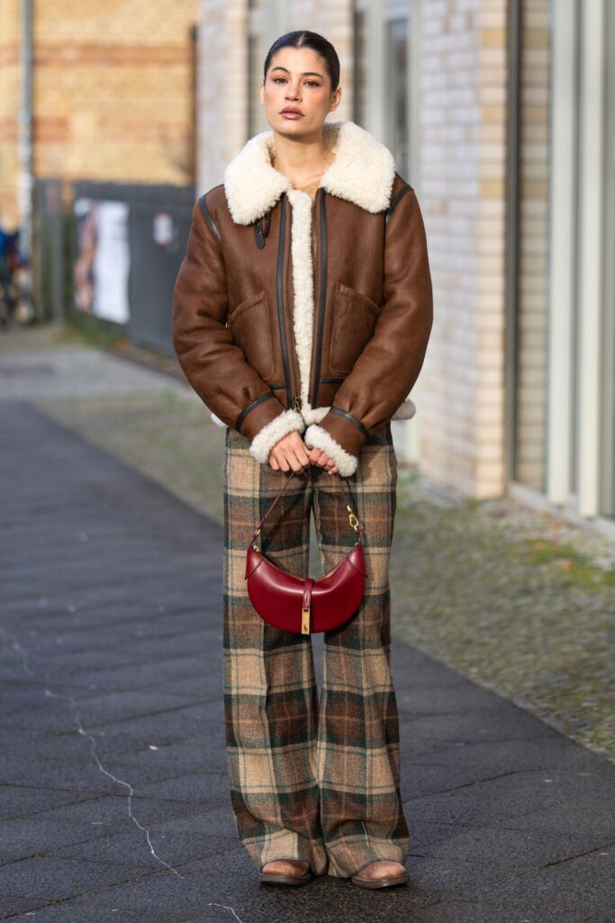 BERLIN, GERMANY - NOVEMBER 26: Lea Naumann wears brown white shearling jacket, beige pullover & red bag Polo Ralph Lauren, brown beige checkered pants Sezane, boots Miu Miu on November 26, 2024 in Berlin, Germany. 